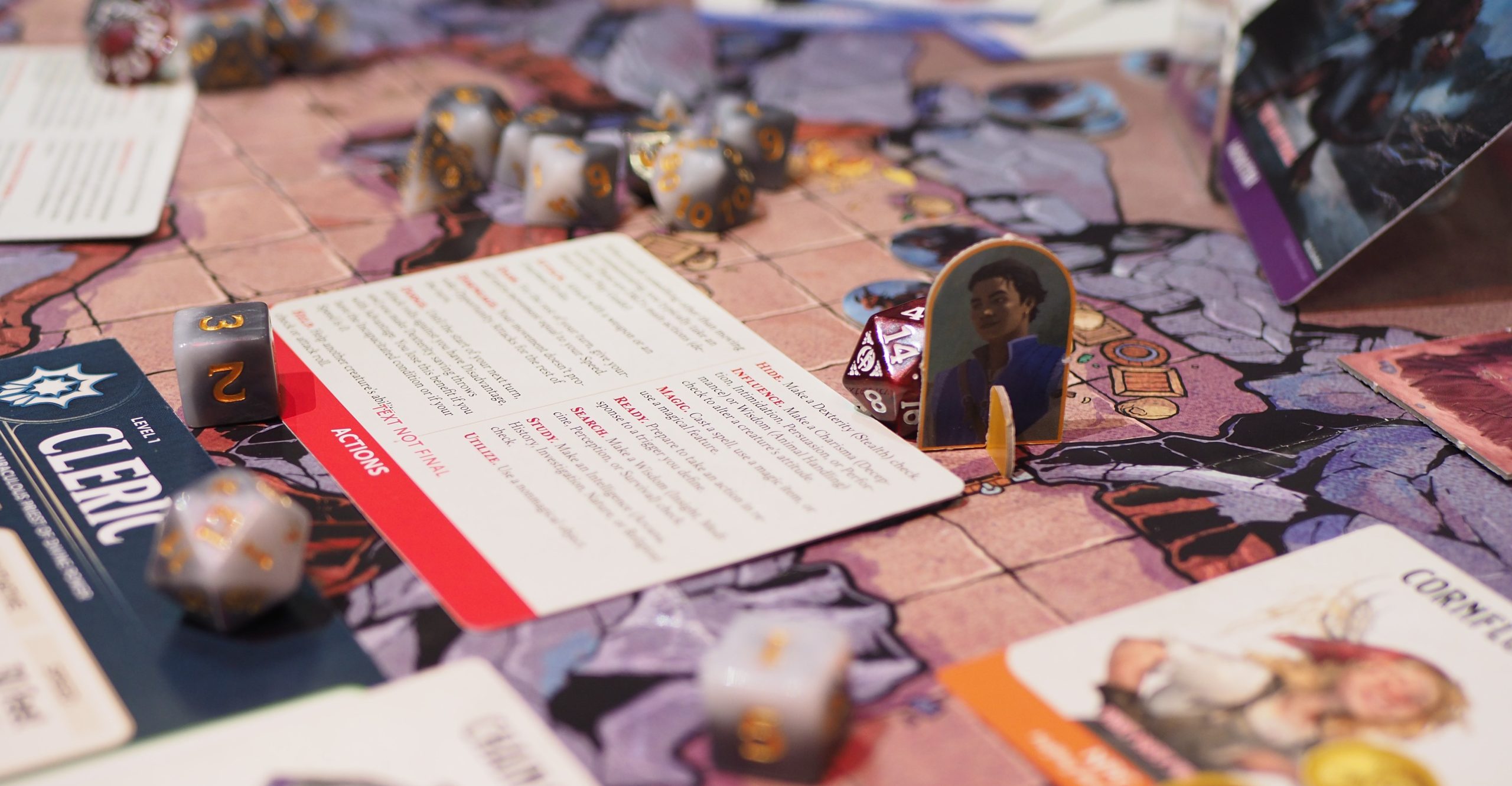 A side angle view of the Caves of Chaos adventure map included with the 2025 Dungeons & Dragons Starter Set. It shows multiple game pieces and a Cleric character sheet, including actions and dice.